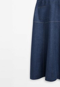 Jupe en jean bleu foncé avec des coutures visibles et une poche arrière rectangulaire, suspendue sur un fond blanc.