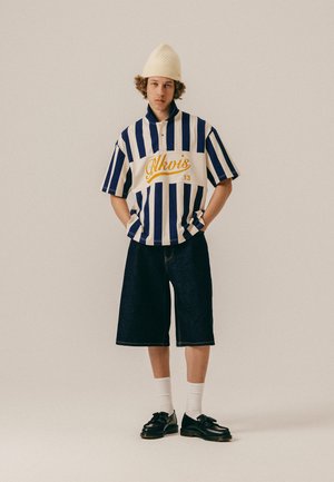 Jauns vīrietis, tērpies zils baltās joslās pārģērbts krekls ar īsām piedurknēm un uzrakstu "Alkvis", tumšās garās šortās, baltās zeķēs, melnās kurpēs un bēšā adītā cepurē.