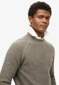 Pull en maille texturée marron avec un col haut, porté avec une chemise blanche en dessous et un petit détail argenté sur la manche.