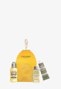 Tre L'OCCITANE-hudvårdsprodukter förpackade i en gul, oval låda. Innehåller en klar duscholja, en enkel oljeflaska och en grön handkräm.
