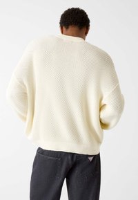 Beiger Strickpullover mit offenem Muster und weiten Ärmeln, kombiniert mit dunkelgrauen Jeans, die aufgesetzte Gesäßtaschen und einen kleinen Logopatch haben.