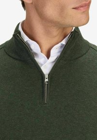 Mörkgrön zip-up tröja gjord av ett texturerat tyg. Har en hög krage och silverdragkedja, lager på lager över en vit skjorta.