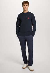 Marineblå strikkegenser med en sirkulær rød logo, rund hals og ribbestrikkede mansjetter. Kombinert med marineblå bukser og hvite sneakers. Modellen står med hendene i lommene.