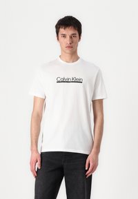 Camiseta blanca de algodón con cuello redondo, que presenta un logotipo negro de "Calvin Klein" centrado en el pecho. Manga corta y corte relajado.