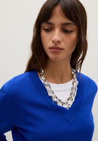 Blauwe v-halstrui van zacht materiaal, gelaagd over een wit shirt, gecombineerd met een robuuste zilveren ketting.