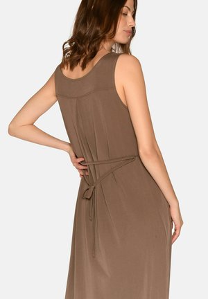 Vrouw draagt een mouwloze bruine jurk met een strikceintuur aan de achterkant, poseert met één hand op haar heup en draait lichtjes opzij.