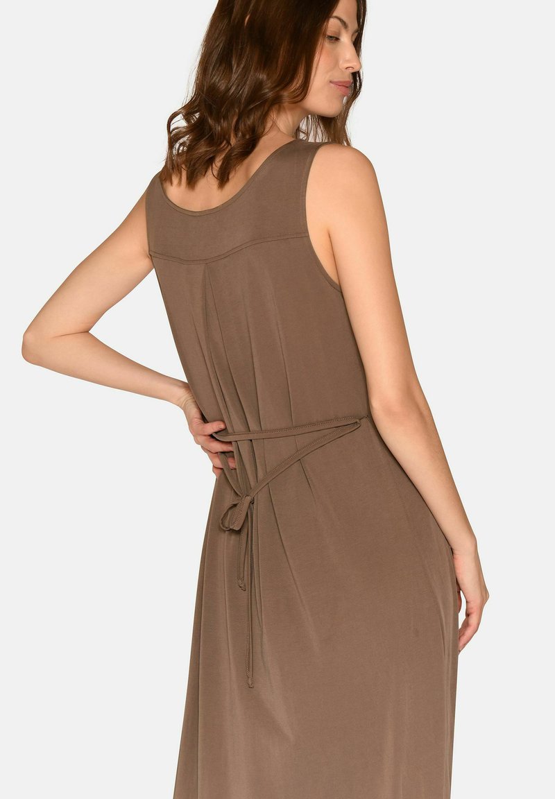 Moteris vilki rudą rankovių neturintį suknelę su juostos diržu, surištu nugaroje, pozuoja viena ranka ant juosmens, šiek tiek pasisukusi šonu.