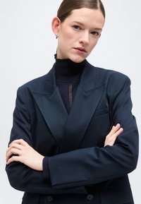 Jeune femme aux cheveux lissés en arrière portant un blazer sombre sur un col roulé noir, debout les bras croisés devant un fond uni.