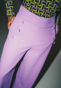 Pantaloni a vita alta color lavanda con tasche laterali, caratterizzati da quattro bottoni rotondi rivestiti in tessuto e una texture liscia.