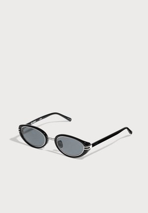 Lunettes de soleil ovales noires avec un pont en métal fin et des verres teintés foncés sur un fond clair.