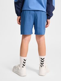 Blå sportshorts i lättviktsmaterial med elastisk midja. Bärs med vita strumpor prydda med svarta Chevron-mönster och träningsskor.