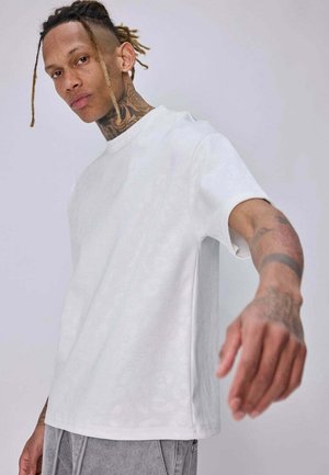 Jeune homme avec des dreadlocks et des tatouages, portant un t-shirt blanc uni et un pantalon gris, tend la main vers la caméra sur un fond uni.