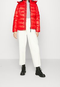 Röd vadderad jacka med huva, kombinerad med en grå topp och vita vida byxor. Svarta snörade kängor fulländar outfiten.