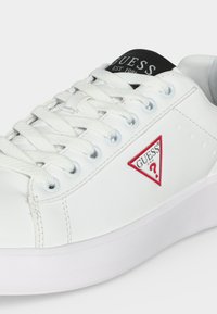 Άσπρο sneaker Guess με λευκά κορδόνια, μαύρη γλώσσα με την επιγραφή "Guess EST. 1981" και κόκκινο τριγωνικό λογότυπο Guess στο πλάι.