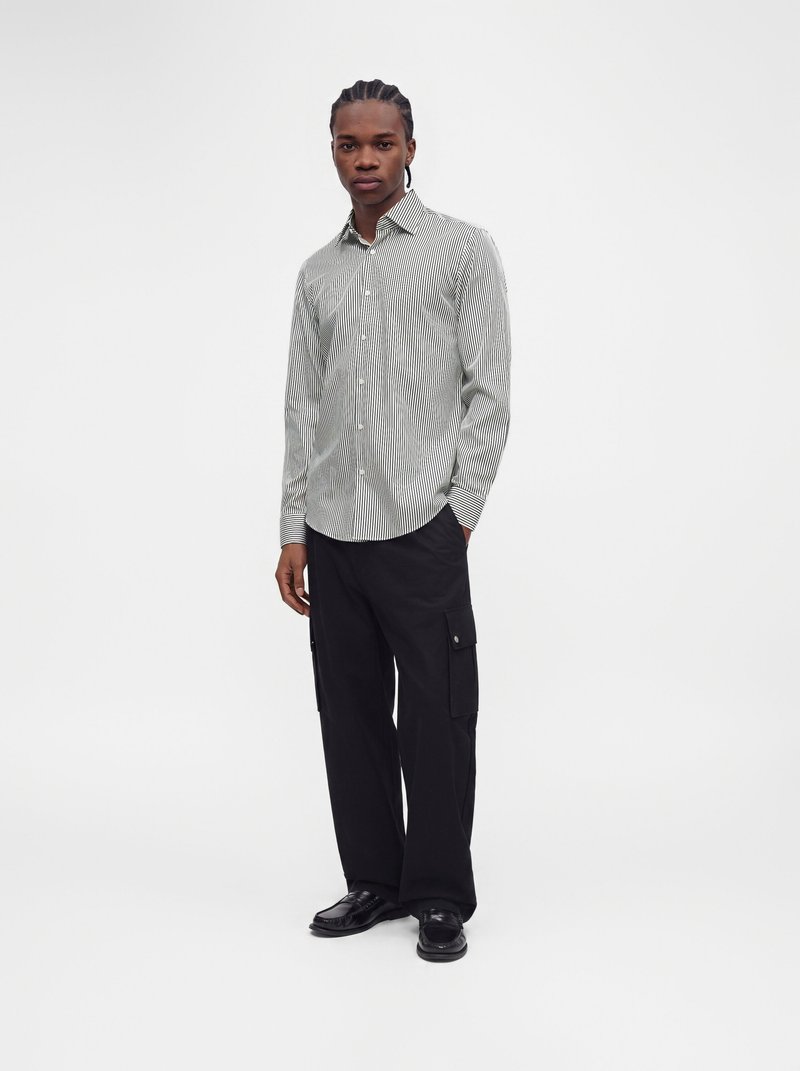 Homme debout portant une chemise fine à rayures à manches longues, un pantalon cargo noir et des chaussures noires sur un fond blanc uni.