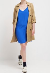 Gabardina beige con mangas remangadas sobre un vestido sin mangas azul. El conjunto se complementa con zapatos blancos y calcetines oscuros.