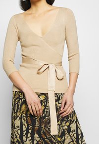 Beige randig sveptopp med djup V-ringning, trekvartslånga ärmar och ett knytband i midjan, tillsammans med en mönstrad svart kjol.