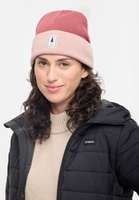 Gorro de lana en rosa y granate con una etiqueta de logo bordada. El modelo lleva una chaqueta acolchada negra y un suéter beige de canalé por debajo.