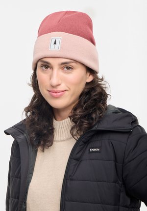 Knitted beanie in pink and maroon with a stitched logo label. Model wears a black puffer jacket and a beige, ribbed sweater underneath.