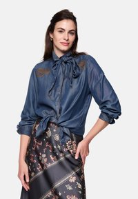 Denim shirt with bow tie, featuring embroidered accents on the shoulders. Paired with a patterned, long, dark skirt.