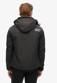 Zwarte capuchonjack met een gestructureerd oppervlak en subtiel ruitpatroon. Heeft een logo op de achterkant en elastische manchetten voor een snug fit.