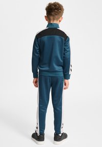 Teal trainingspak met zwarte accenten, bestaande uit een zipperjack en bijpassende broek. Gestructureerde stof en contrasterende zijstrepen. Zwarte sneakers.