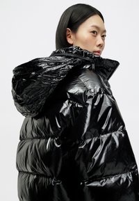 Μαύρο μπουφάν με γυαλιστερή επιφάνεια, με κουκούλα, κλείσιμο με φερμουάρ και οριζόντιο ραμμένο σχέδιο που δημιουργεί μια αφράτη υφή.