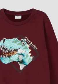 Bordowy sweter z okrągłym dekoltem, ozdobiony wielokolorowym wzorem T-Rexa z cekinami oraz napisem "T-REX NATION". Miękka tekstura.