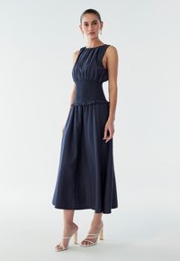 Vestido sin mangas azul marino con cintura fruncida y falda acampanada. Tela texturizada, dobladillo con volantes, combinado con tacones blancos de tiras.