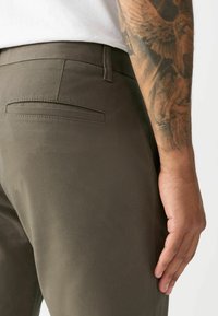 Olive green trousers with a smooth texture and a single back pocket; hand resting on the side, featuring a visible tattoo.