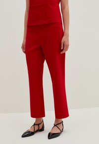 Person wearing red tailored ankle-length pants, red top, and black patent leather pointed flats with ankle straps, standing on a light background.
