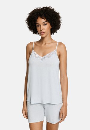 Donna con capelli ricci che indossa una canottiera azzurra con pizzo e pantaloncini coordinati, in piedi davanti a uno sfondo neutro.