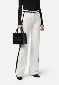 Pantaloni bianchi a gamba larga con strisce nere sui lati, abbinati a una borsa nera con logo testurizzato e scarpe nere con punta appuntita.