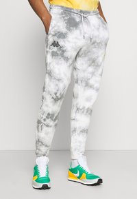 Grå batikmönstrade sweatpants med elastisk midja, sidofickor och en svart logotyp. Bärs med gröna och gula sneakers, med vita snörningar.