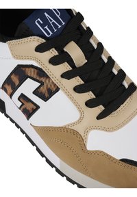 Gros plan sur une basket avec des panneaux beige et blanc, des lacets noirs, un logo "G" à motif léopard, et une étiquette bleue avec le texte "GAP".