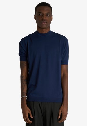 Giovane uomo con capelli corti e orecchini indossa una maglietta blu navy a maniche corte tinta unita e pantaloni neri, in piedi davanti a uno sfondo bianco.