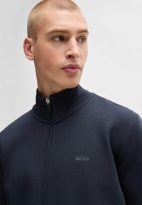 Marinblå zip-up jacka med hög krage tillverkad av slätt tyg. Har en diskret logga på bröstet och en snygg, modern design.