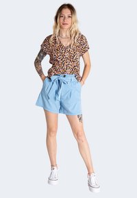 Haut à manches courtes avec un motif floral en noir, orange et jaune ; associé à un short taille haute bleu clair avec une ceinture nouée. Baskets blanches.