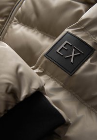 Beige polstret jakke med en sort stofmanchet og et firkantet logo med "EX" i forhøjet skrift på ærmet. Glat, skinnende tekstur.