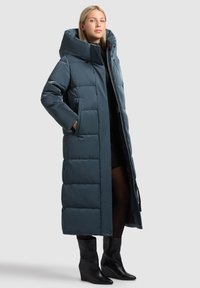 Pitkä, tummansinivihreä untuvatakki, jossa on suuri huppu, vetoketjulla edessä ja kaksi sivutaskua. Sileä, kiiltävä pinta ja tikattu kuvio.