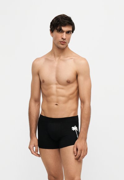Homme torse nu aux cheveux courts et foncés portant un boxer noir avec un motif de palmier blanc sur un fond clair uni.