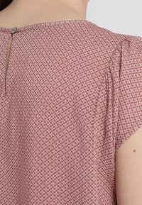Top a maniche corte in rosa pallido con un motivo geometrico bianco. Presenta un'apertura sul retro chiusa da un bottone. Tessuto dalla texture liscia.