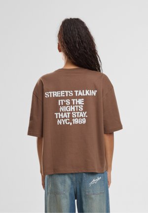 Personne aux longs cheveux bouclés porte un T-shirt marron oversize avec le texte blanc « STREETS TALKIN’ IT'S THE NIGHTS THAT STAY. NYC, 1989 » au dos.