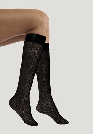 Benendragende zwarte kanten kniekousen met effen zwarte elastische bovenkanten tegen een lichtgrijze achtergrond.