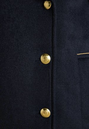 Marineblauer Wollmantel mit fünf goldenen Knöpfen, ausgestattet mit einem strukturierten Stoff und einem subtilen bestickten Detail in der Nähe der Tasche.