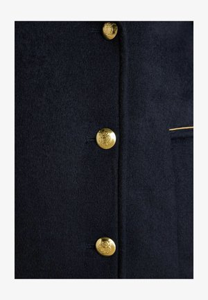 Marineblauer Wollmantel mit fünf goldenen Knöpfen, ausgestattet mit einem strukturierten Stoff und einem subtilen bestickten Detail in der Nähe der Tasche.