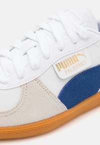 Vita Puma Palermo sneakers med läder- och mockadetaljer i grått och blått, med vita snörningar och en gul logotavla på sidan.