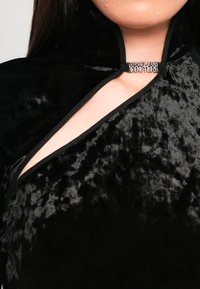 Juodas aksominis palaidinė su aukštu apykaklės ir atviru dekolte, turinti sidabrinį „Versace Jeans Couture“ logotipą užsegime ir tekstūruotą paviršių.