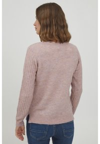 Maglione rosa chiaro con maniche a costine, morbida texture, scollatura rotonda e fenditure laterali; abbinato a jeans blu scuro.