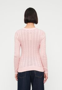 Femeie cu păr scurt și închis la culoare, purtând un pulover roz deschis cu model împletit și blugi albaștri închiși, stând cu fața spre spate pe un fundal simplu.